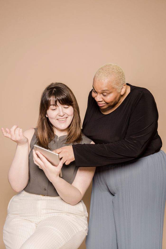 Positive plus size multiracial female coworkers surfing internet on modern tablet while standing on beige background in light studio during work together