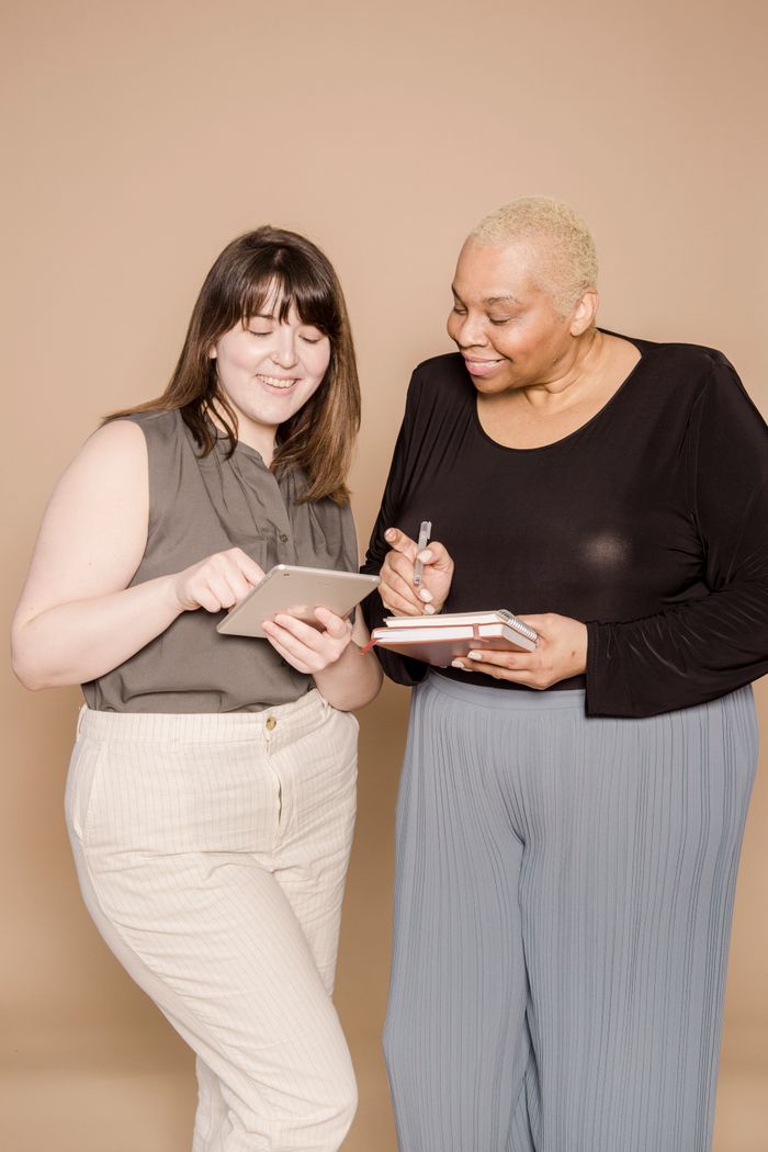 Two women, one holding a notebook and pen, smiling while collaborating together indoors.