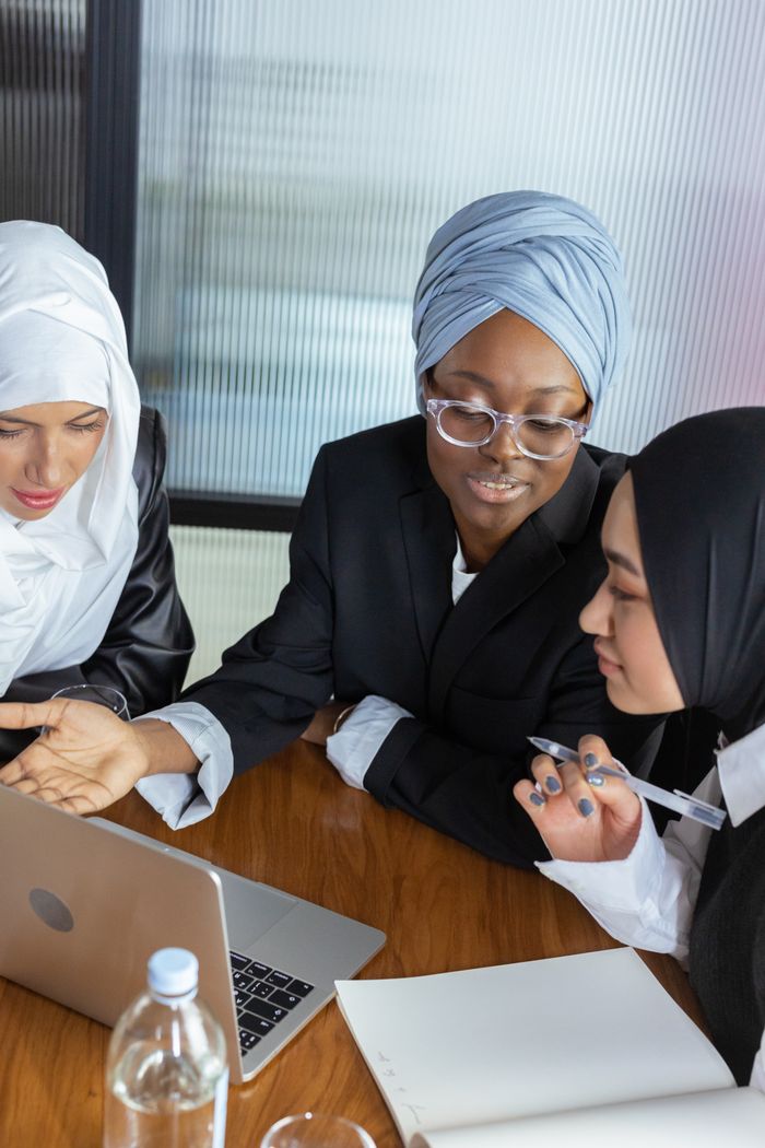 hero-contact Three multicultural businesswomen in hijabs discuss work around a laptop in an office setting.
